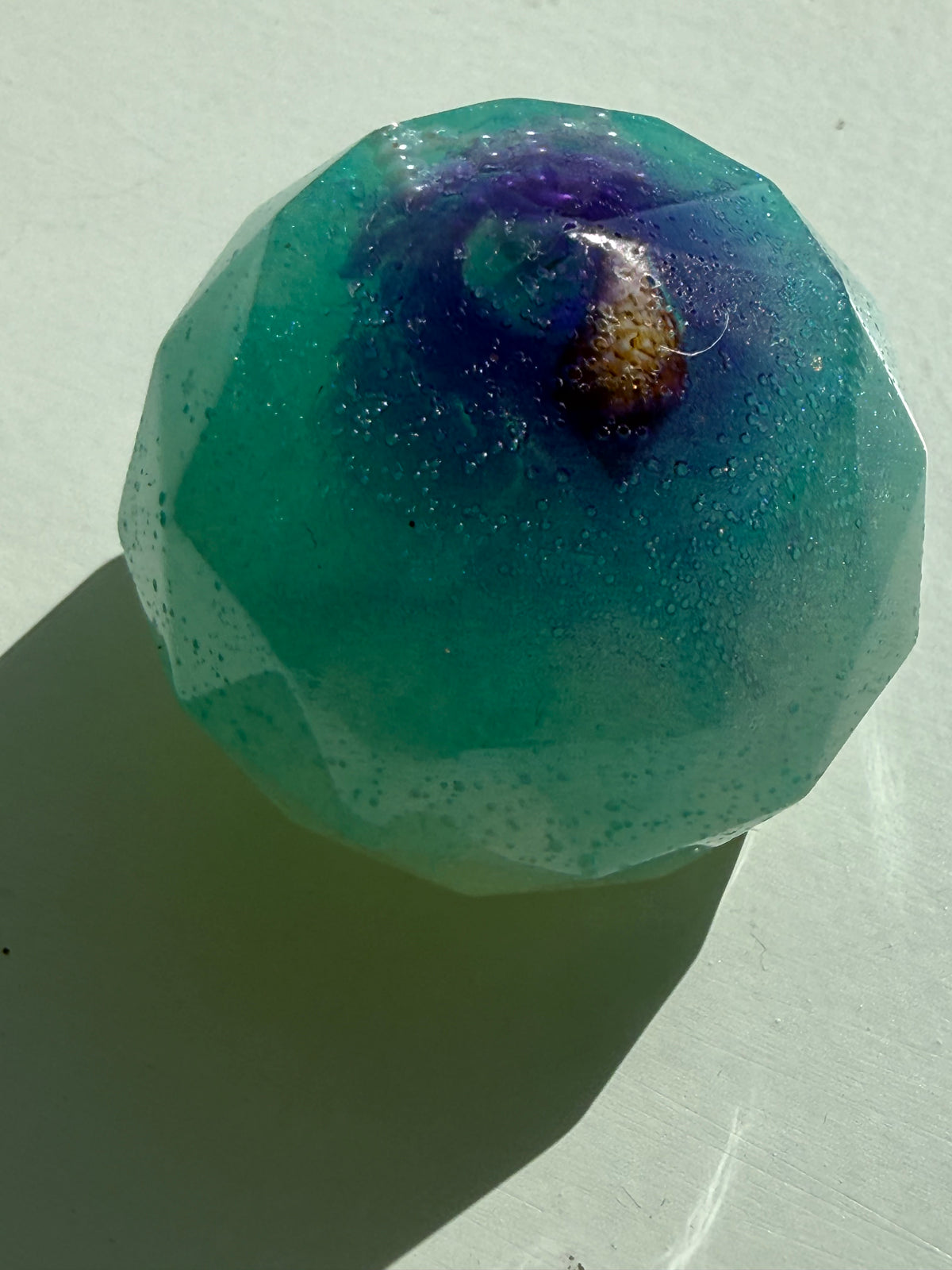 A faceted, translucent teal resin orb with embedded glitter and a dark purple swirl near the top, resting on a light-colored surface.